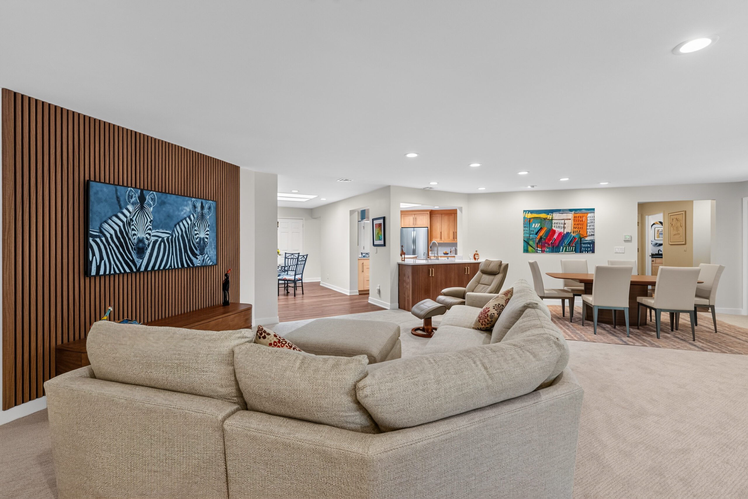 Open-concept living area with beige sectional sofa facing a wooden slatted wall and zebra artwork mounted on it, spacious and bright.