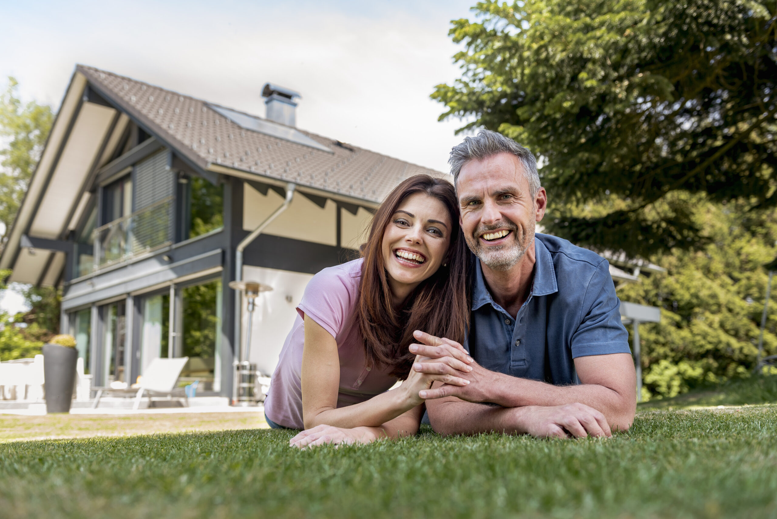 Smiling couple lying on the grass in a backyard with a modern house and trees in the background.