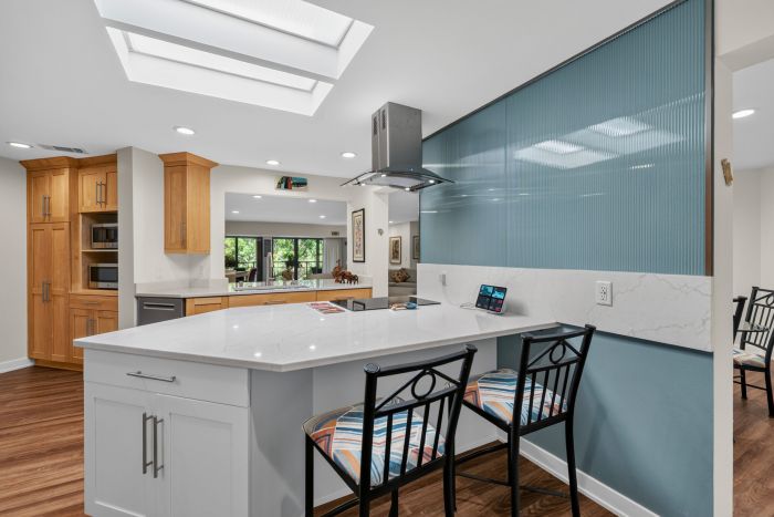 Bright open kitchen with a white island, warm wooden cabinets, skylights, and a blue ribbed glass divider on the right.