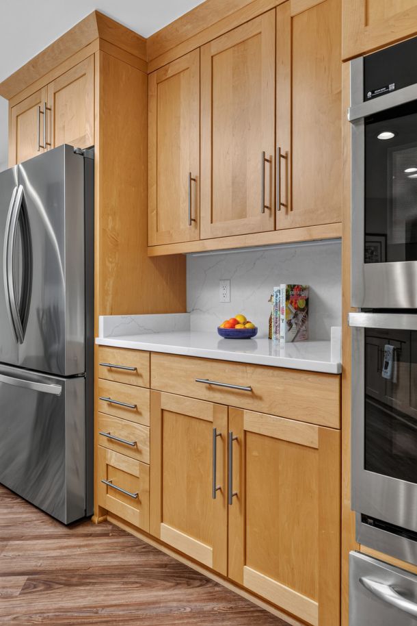 Light wood kitchen cabinets with stainless steel handles above a white marble countertop and a bowl of fruit on the counter