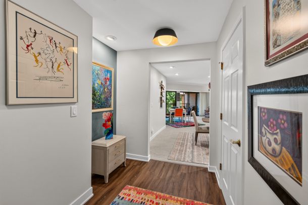 Bright hallway with framed abstract and floral artwork on light gray walls, leading to a living/dining area with colorful chairs and large windows in the distance.