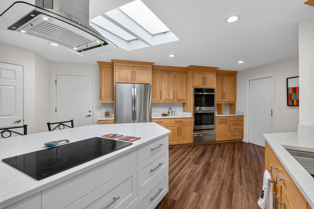Modern kitchen with white countertops and island, stainless steel fridge and double ovens, and warm wood cabinets under a skylit ceiling.