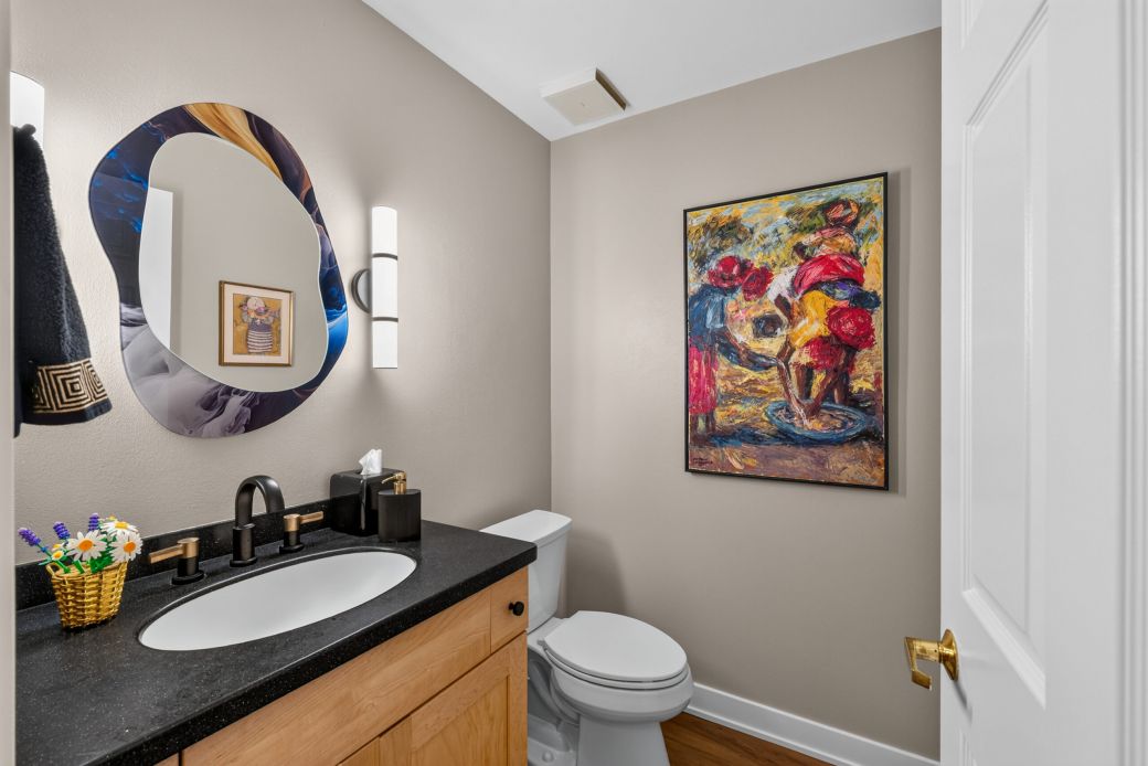 Bathroom with a two-sink vanity, round multicolored mirror, and abstract painting on beige walls; tissue box and small flower basket on counter, toilet nearby.