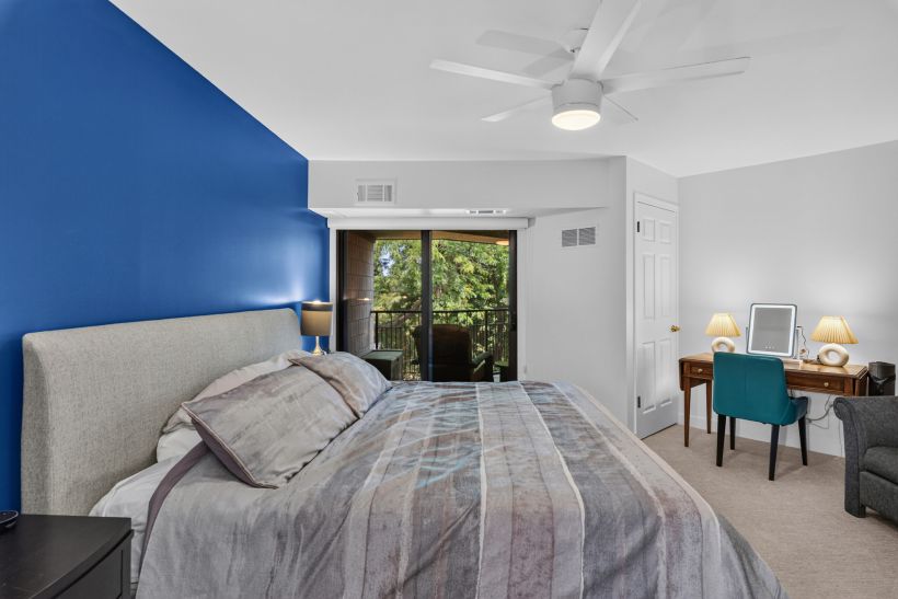 Blue accent wall behind a grey upholstered bed; a balcony door and desk with lamps visible to the right.