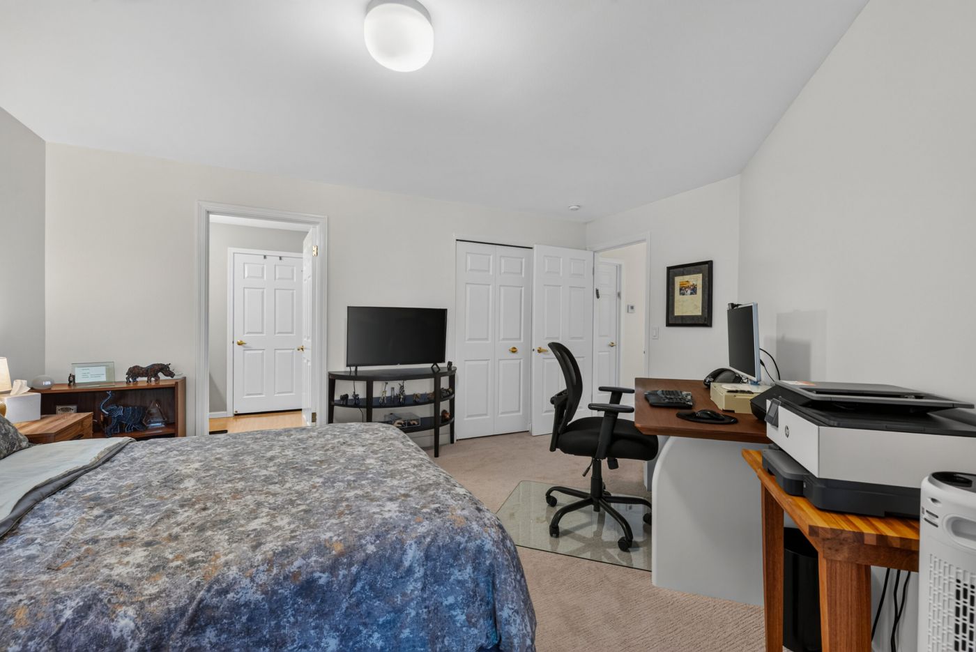 Bedroom with a workspace: bed in the foreground and a desk with computer, printer, and office chair on the right; white walls and doors in the background.