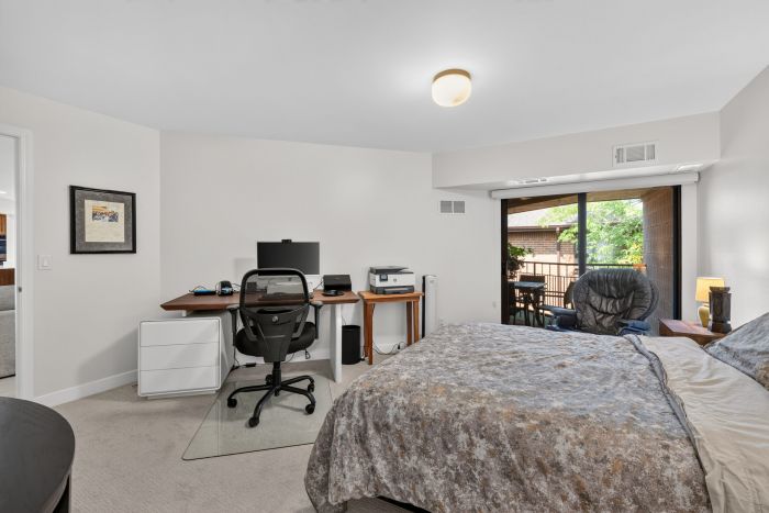 Bedroom with a desk setup and a sliding glass door opening to a balcony; computer monitor, printer, and office chair on the desk, and a large bed with grey patterned bedding in the foreground.