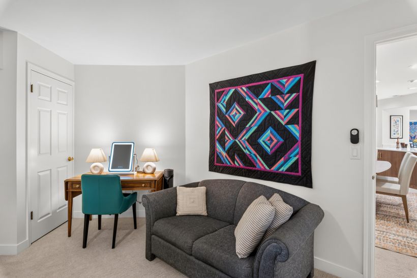 A living room corner with a colorful geometric quilt on the wall, gray sofa, and a wooden desk with a tablet and two lamps nearby.